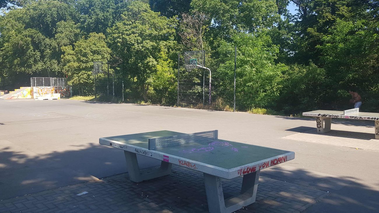 Der Street Court in der Hasenheide: Asphaltplatz mit Basketballkörben und Tischtennisplatten, dahinter Bäume.